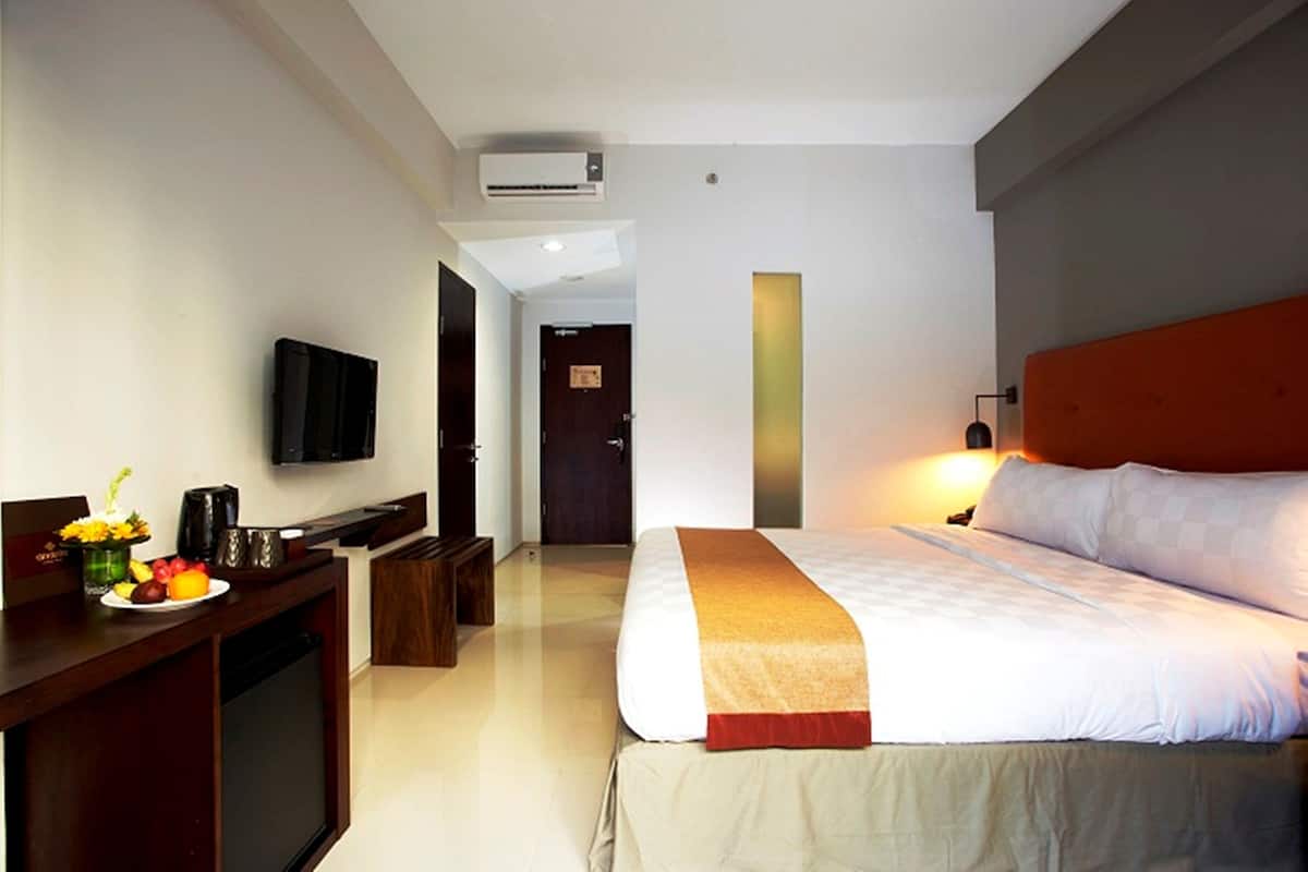 Quarto de hotel com cama, escrivaninha com cafeteira, televisão e ar condicionado fixado na parede.
