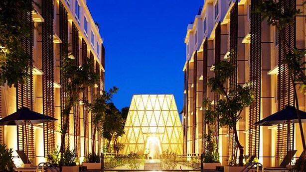 A swimming pool with a geometric glass structure in the background, surrounded by modern buildings with balconies and outdoor seating. - Ananta Legian Hotel (Legian)