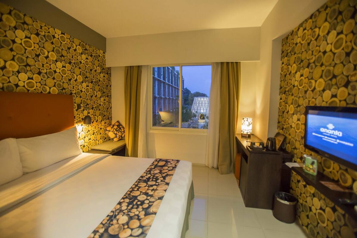 Quarto de hotel com cama, escrivaninha, televisão e janela com cortinas.