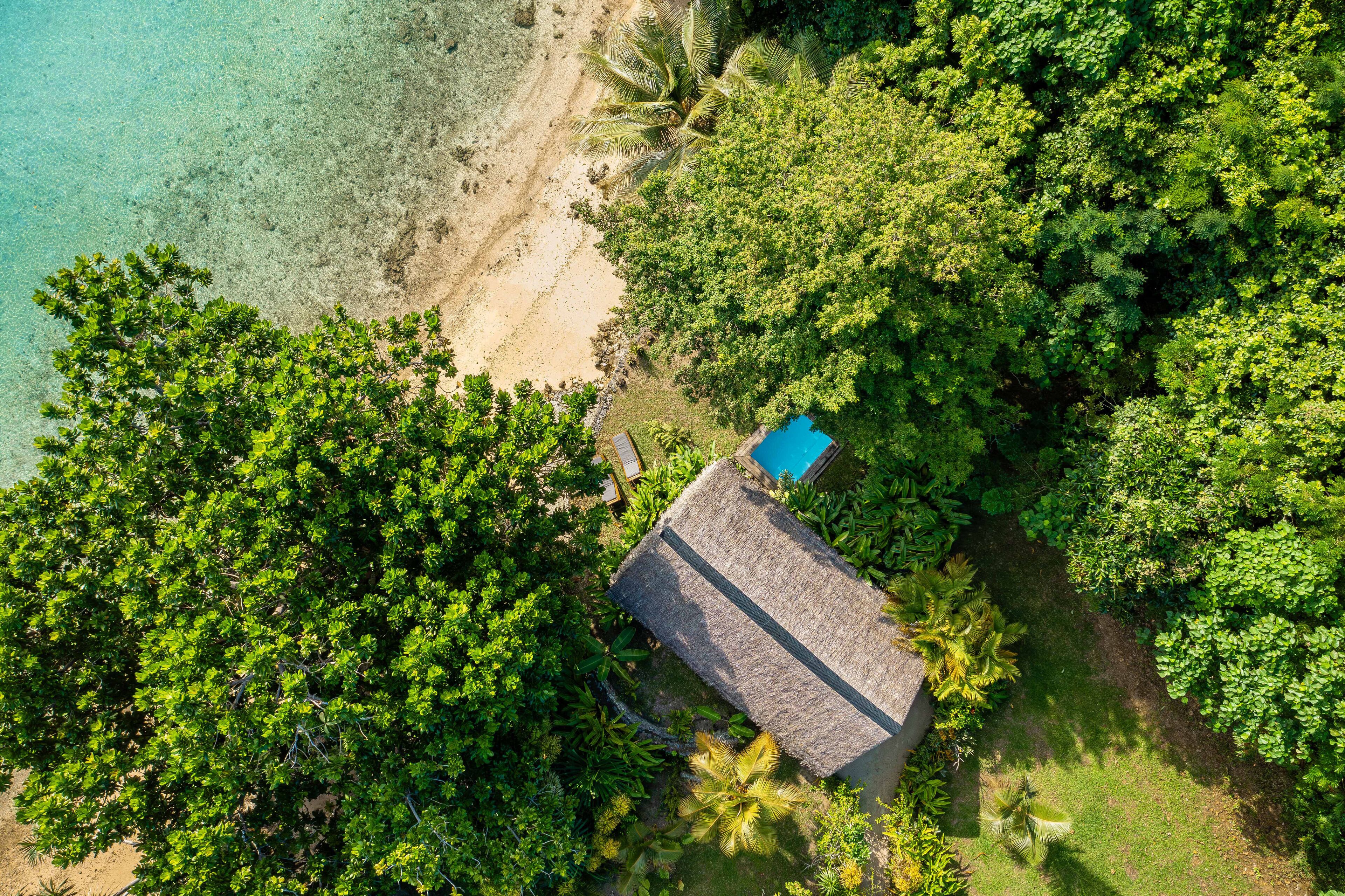 Vista aérea de una casa de playa aislada rodeada de exuberante vegetación tropical, con un océano azul cristalino y una pequeña piscina.