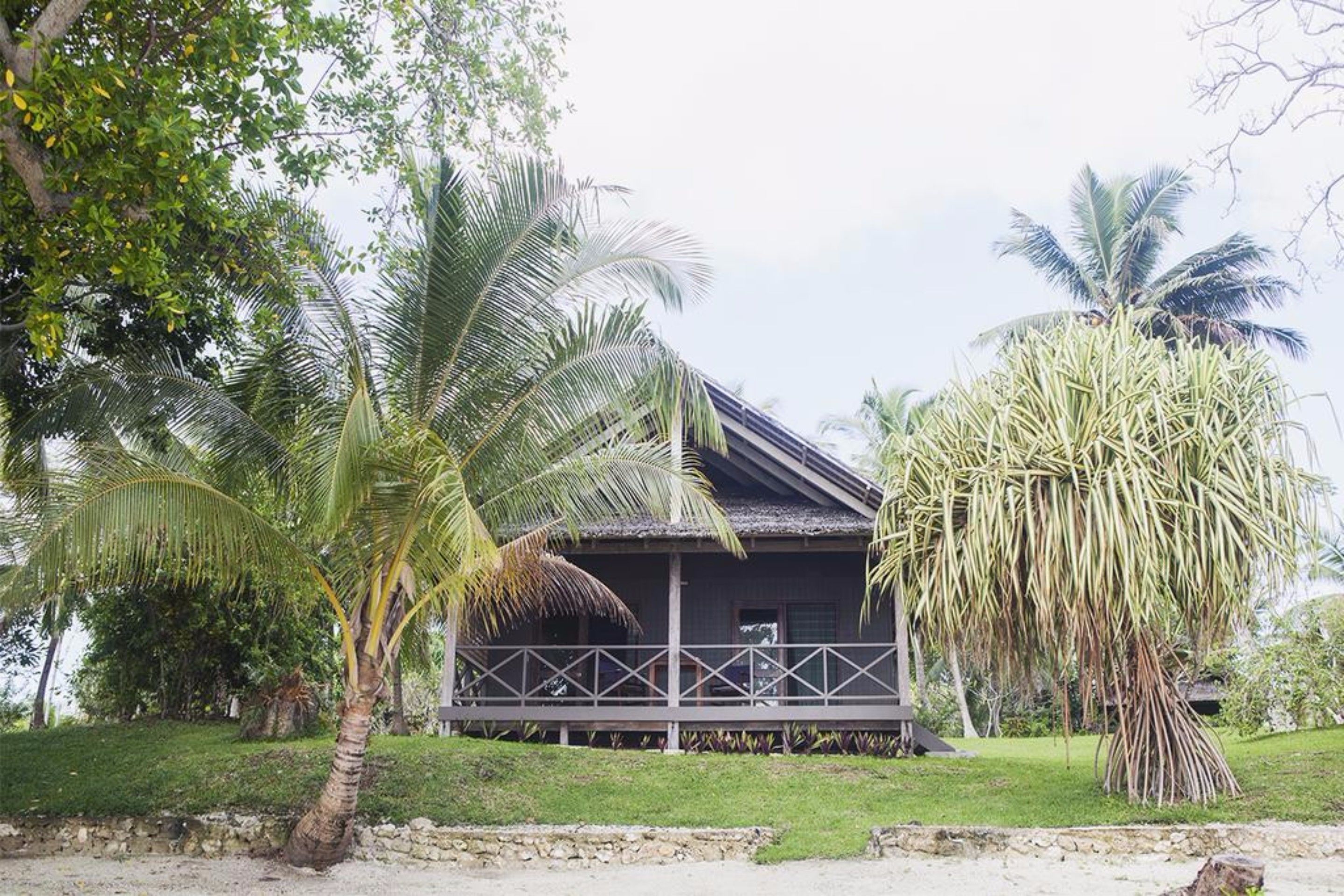 A wooden house with a porch, surrounded by palm trees and a stone wall.