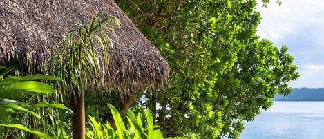 A pool area with a thatched-roof structure, surrounded by lush greenery and overlooking a body of water.