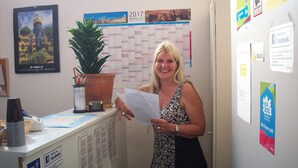 A woman holding a paper in an office with a calendar, a plant, and various notices on the wall. - Gaestehaus Kaiserpassage - Hostel (Karlsruhe)