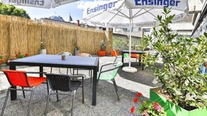 An outdoor seating area with a table and chairs, surrounded by umbrellas with the 'Ensinger' logo, and a backdrop of a straw wall and greenery. - Gaestehaus Kaiserpassage - Hostel (Karlsruhe)