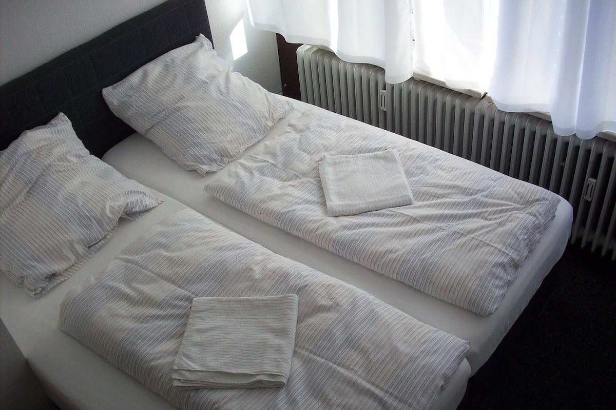 A twin bed with white bedding and pillows, a headboard, and a window with curtains.