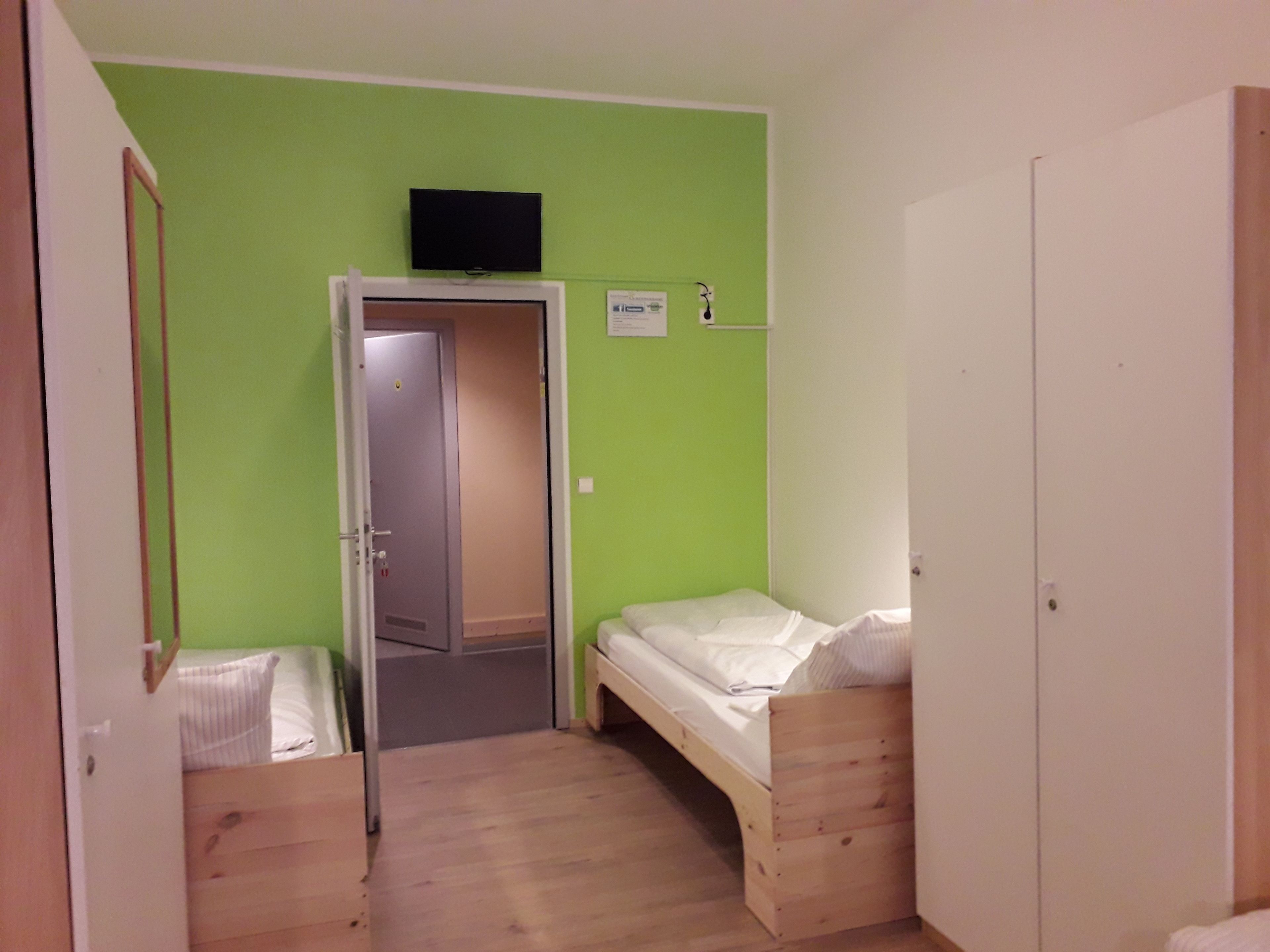 Une petite pièce avec un lit simple, une porte donnant sur une autre pièce et un téléviseur fixé au mur.