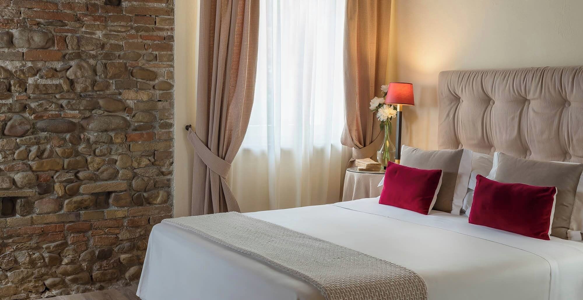 Una habitación de hotel con pared de piedra, cama con ropa blanca y almohadas rojas, cabecero capitonado, mesita de noche con lámpara y cortinas translúcidas.