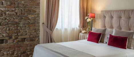 Una habitación de hotel con pared de piedra, cama con ropa blanca y almohadas rojas, cabecero capitonado, mesita de noche con lámpara y cortinas translúcidas.