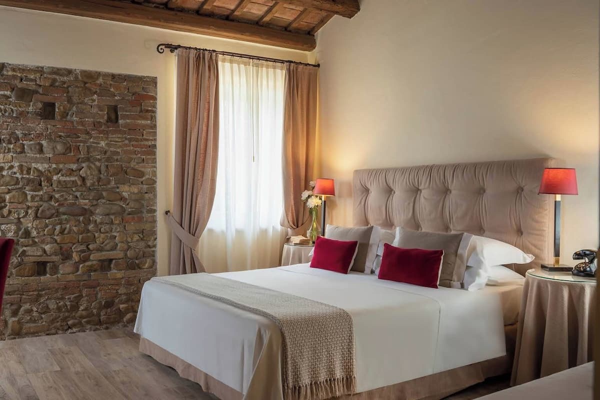 A bedroom with a stone wall, a bed with red pillows, a wooden chair, and a nightstand with a lamp.
