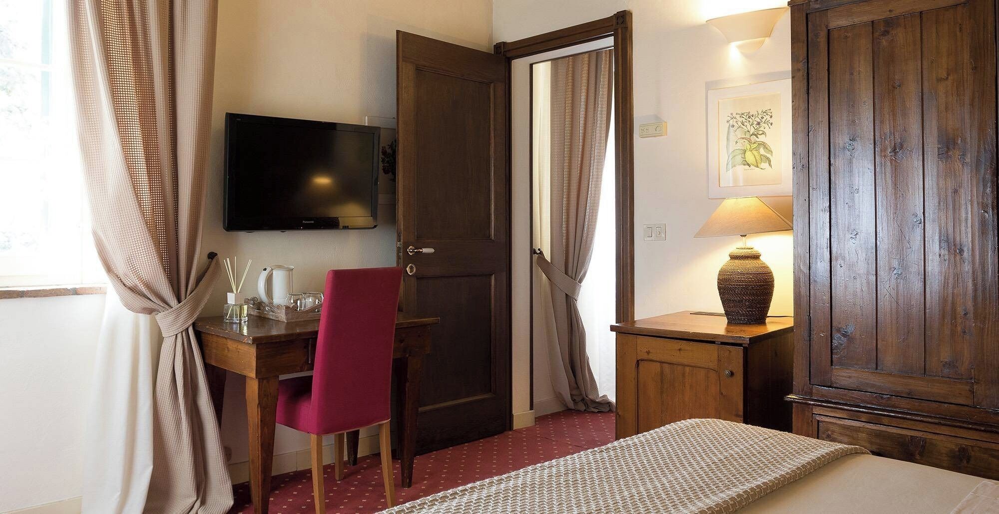 A hotel room with a wooden desk, a red chair, a television, a lamp, and a bed.