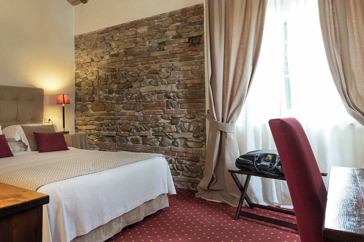 A bedroom with a stone wall, a bed with white bedding and red pillows, a wooden nightstand, a lamp, and a chair with a handbag.