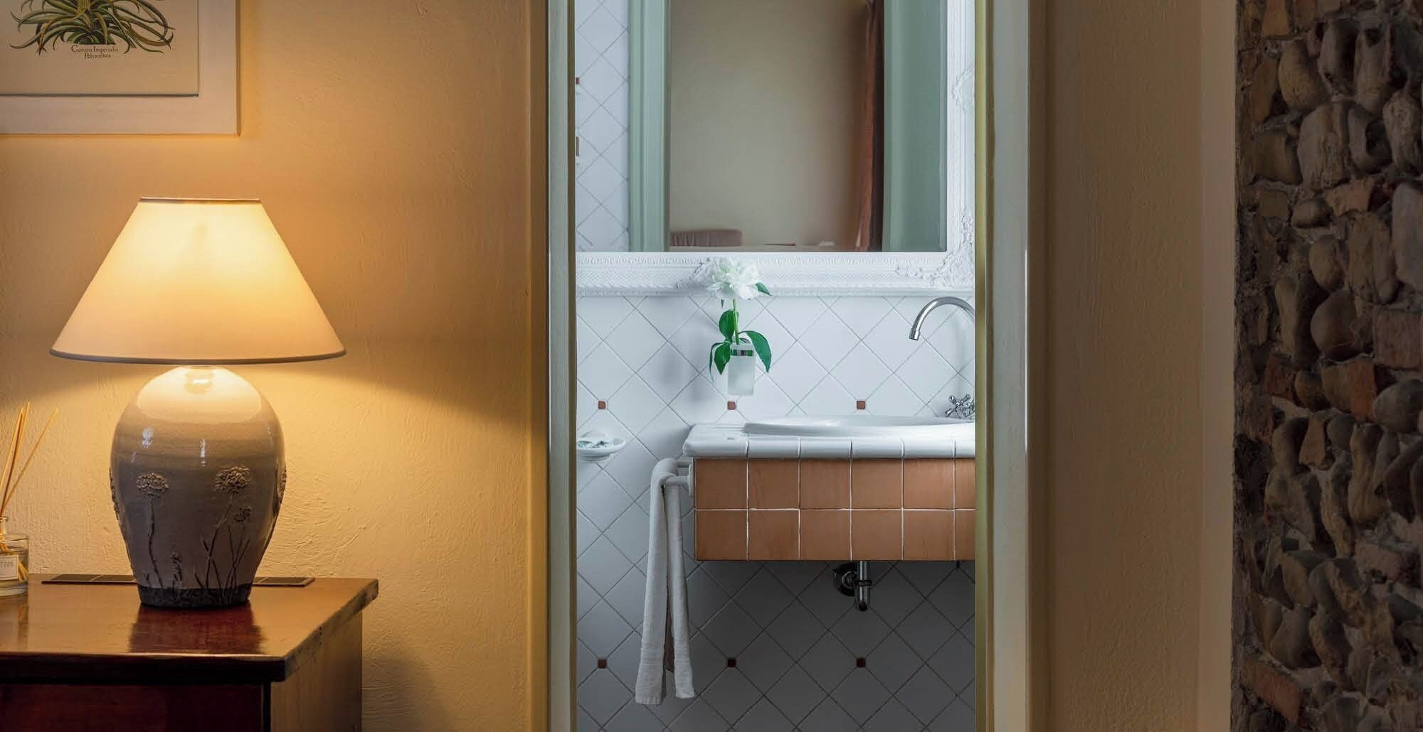 Een badkamer met een witte wastafel, een spiegel, een hangplant en een lamp op een houten bijzettafeltje.