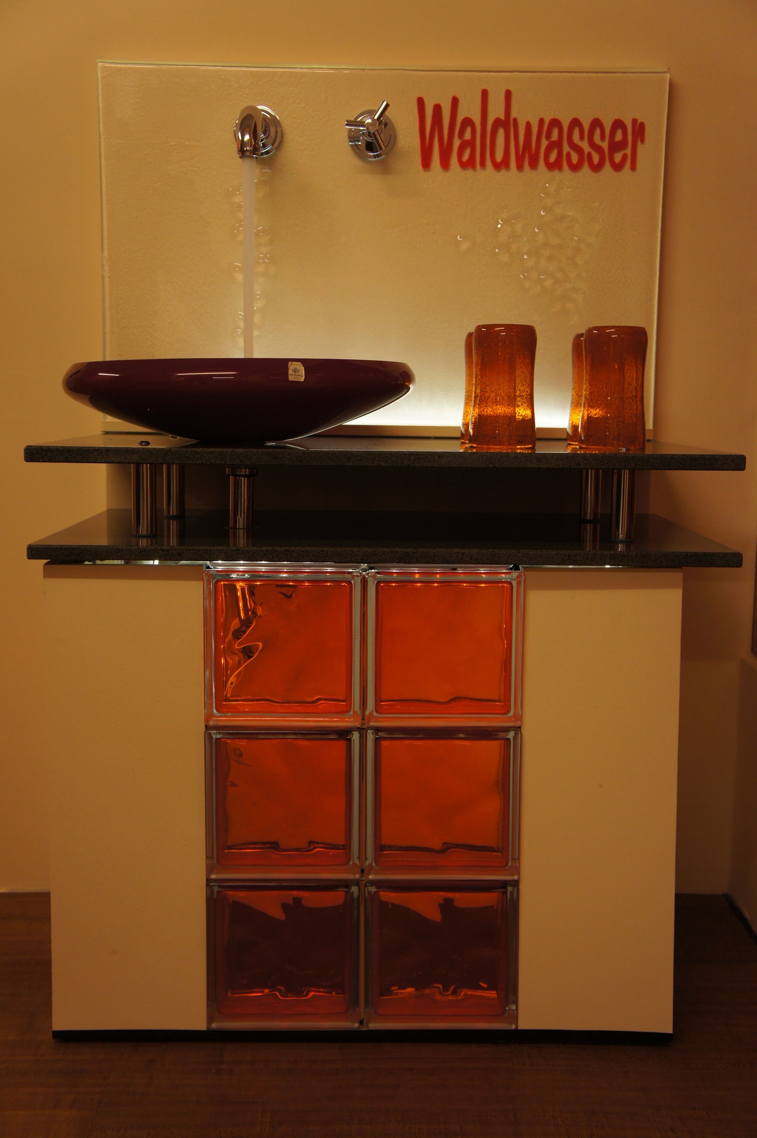 Un baño con un lavabo de vidrio, dos vasos ámbar y un cartel que dice "Waldwasser".