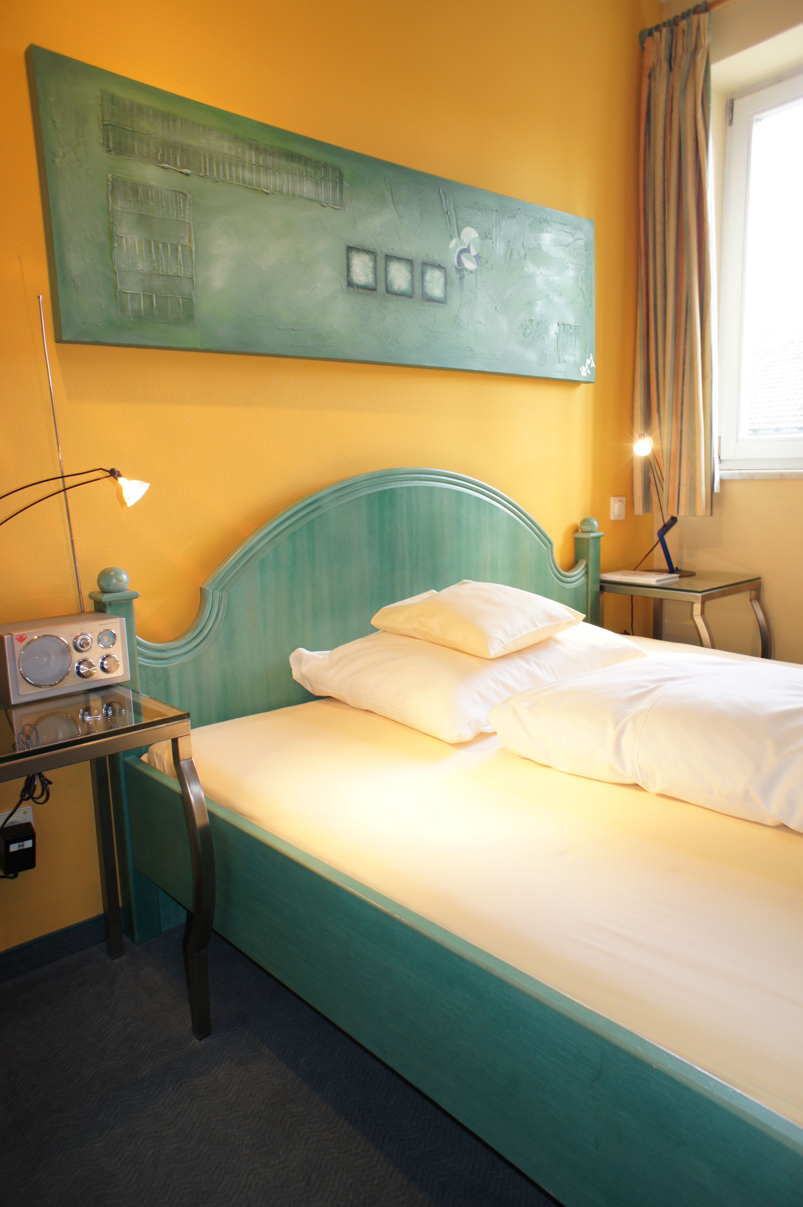 A bed with a teal headboard, a radio on a bedside table, and a wall art piece above the bed.