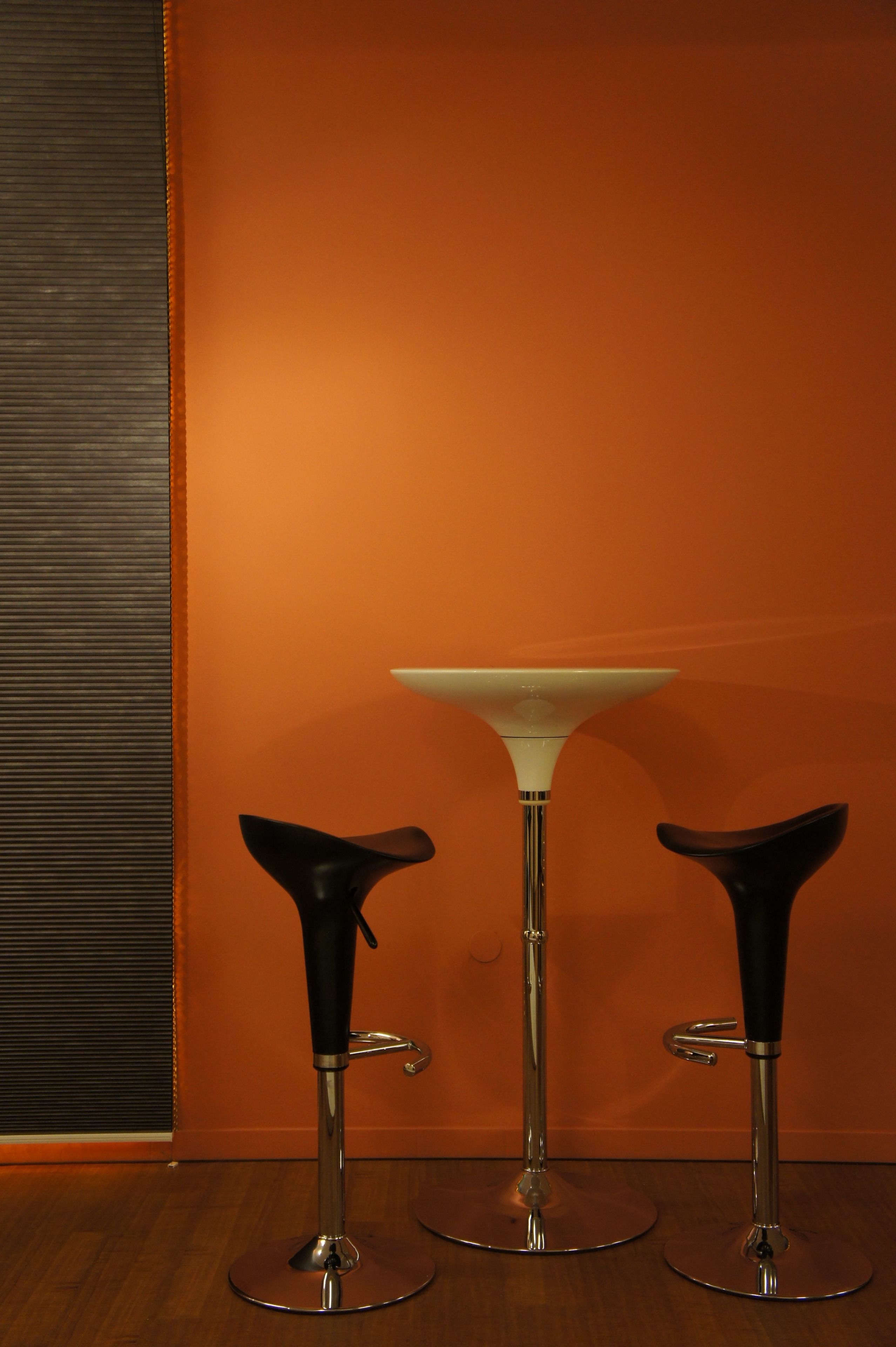 Três banquetas com uma mesa de vidro em um quarto com parede laranja e chão de madeira.
