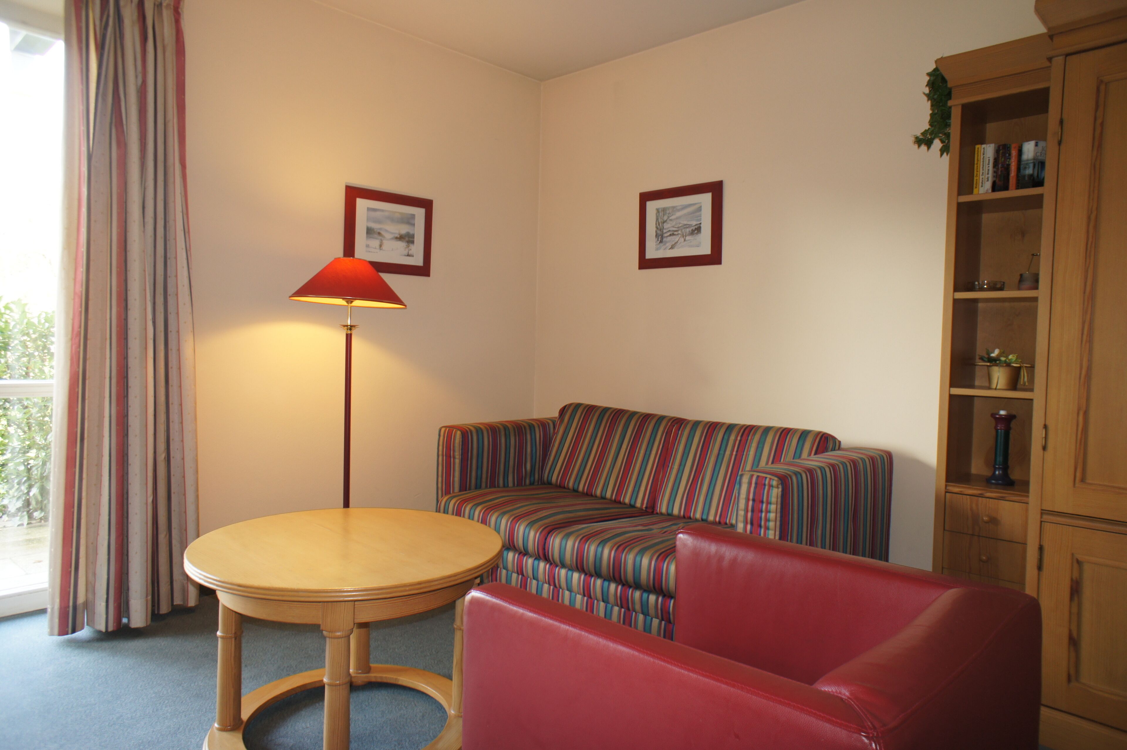 Una habitación con un sofá a rayas, un sillón rojo, una mesa de madera y una lámpara. Hay cuadros en la pared y un aparador con libros.