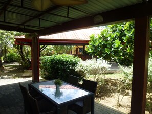 Un patio al aire libre con una mesa de vidrio y sillas de mimbre, rodeado de exuberante vegetación y una estructura de techo rojo al fondo.