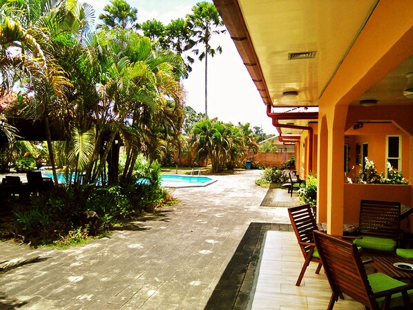 Zona de piscina con un suelo de baldosas, rodeada de exuberante vegetación y palmeras. Hay bancos de madera con cojines verdes a lo largo de la piscina.