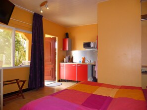 A bedroom with a bed, a window with curtains, a small desk with a chair, and a kitchenette with a microwave.