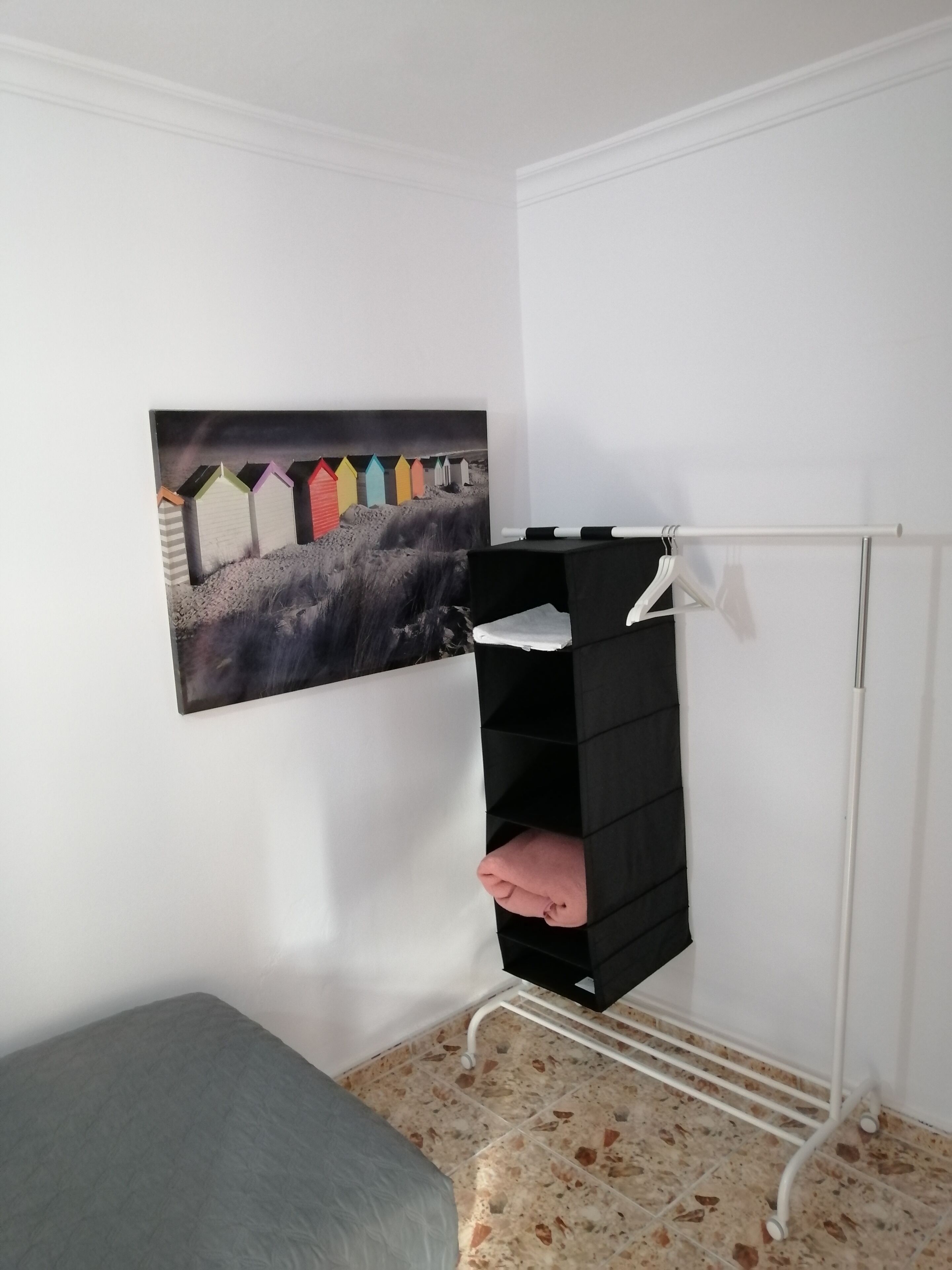 Una habitación con una cama, una pared blanca, un cuadro de cabañas de playa coloridas y un mueble de almacenamiento negro.