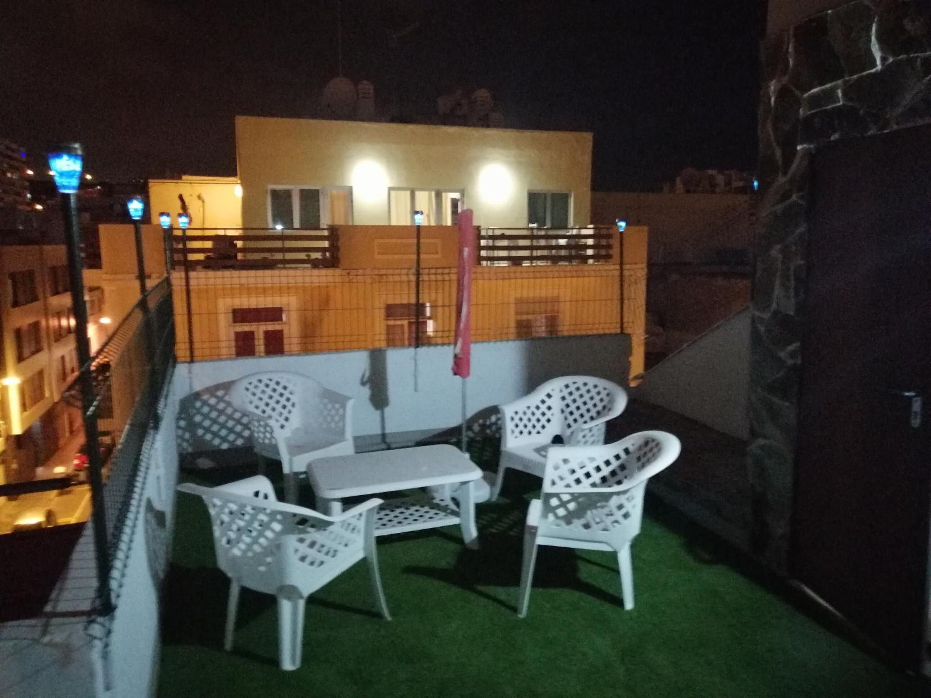 Un balcón con sillas de plástico blanco y una pequeña mesa, un piso de césped artificial verde, y un edificio con balcones al fondo.