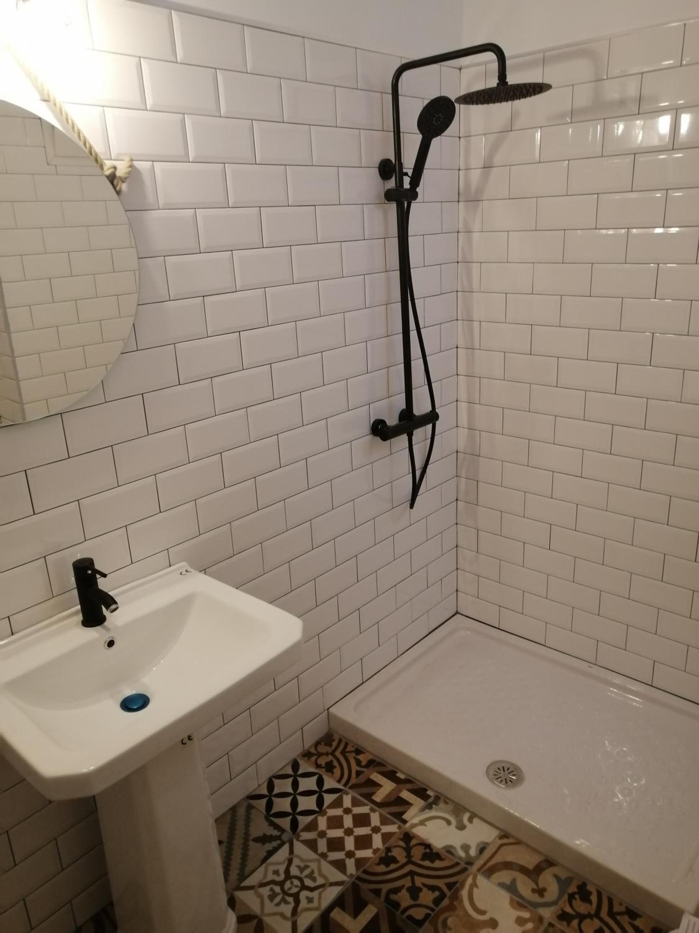 A bathroom with a white sink, a mirror, a shower with a black hose, and patterned floor tiles.