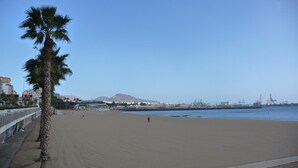 Una amplia playa arenosa con una palmera a la izquierda, cielo azul despejado y edificios industriales al fondo.