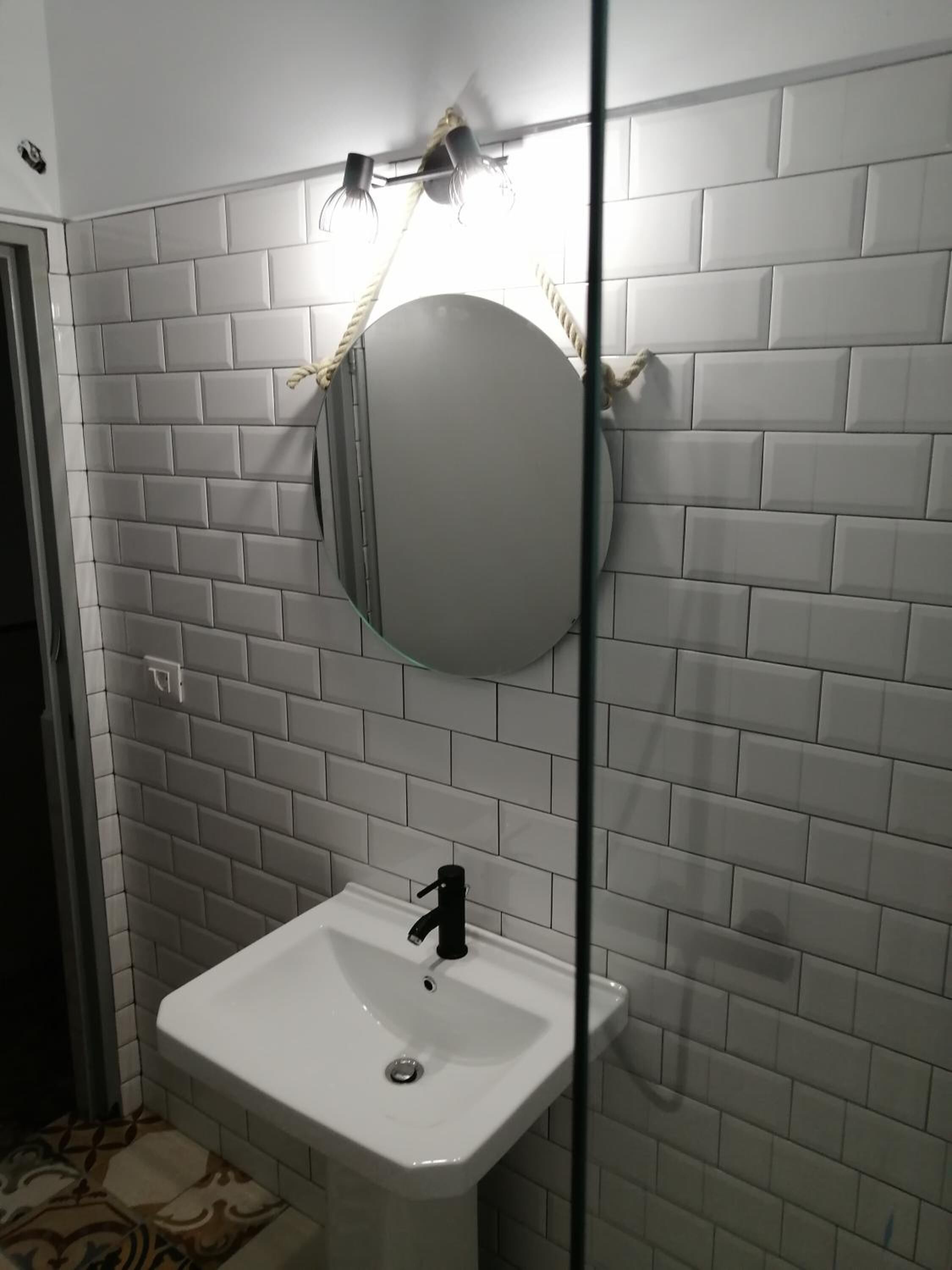 Un bagno con pareti rivestite di piastrelle bianche a metro, uno specchio rotondo e un lavandino bianco con rubinetto nero.