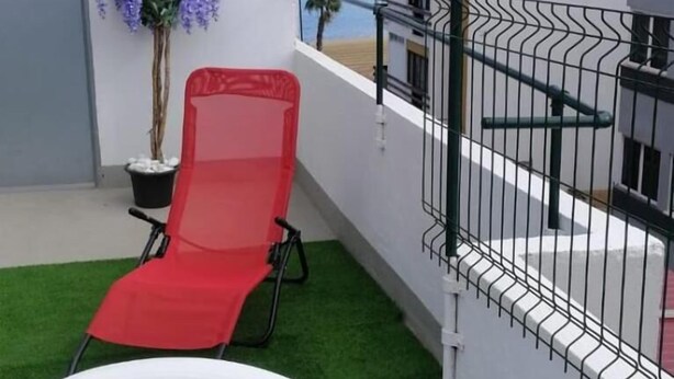 Un balcón con césped artificial, una silla lounge roja, una silla de plástico blanco y una planta en maceta.