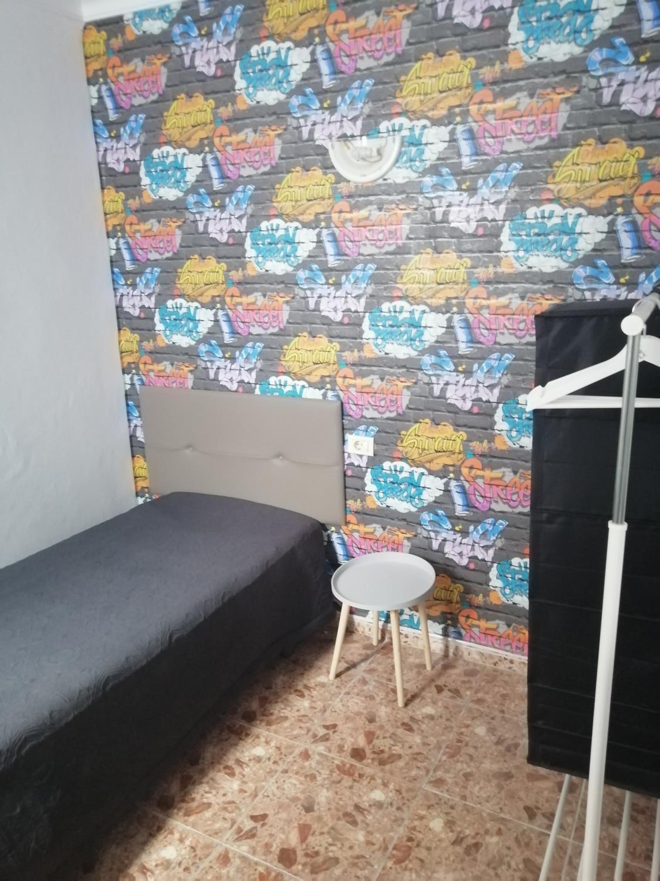 A small room with a single bed, a small table, and a wall covered in colorful graffiti art.