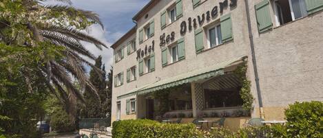 Ein Hotel namens "Hotel des Oliviers" mit einer klassischen Fassade, grünen Fensterläden und einer überdachten Außenbereich-Sitzgelegenheit.