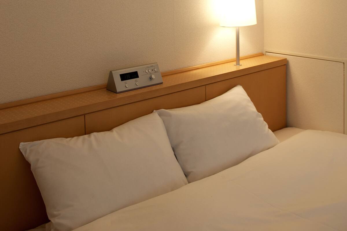 A hotel room with a bed, pillows, a nightstand, and a digital clock.