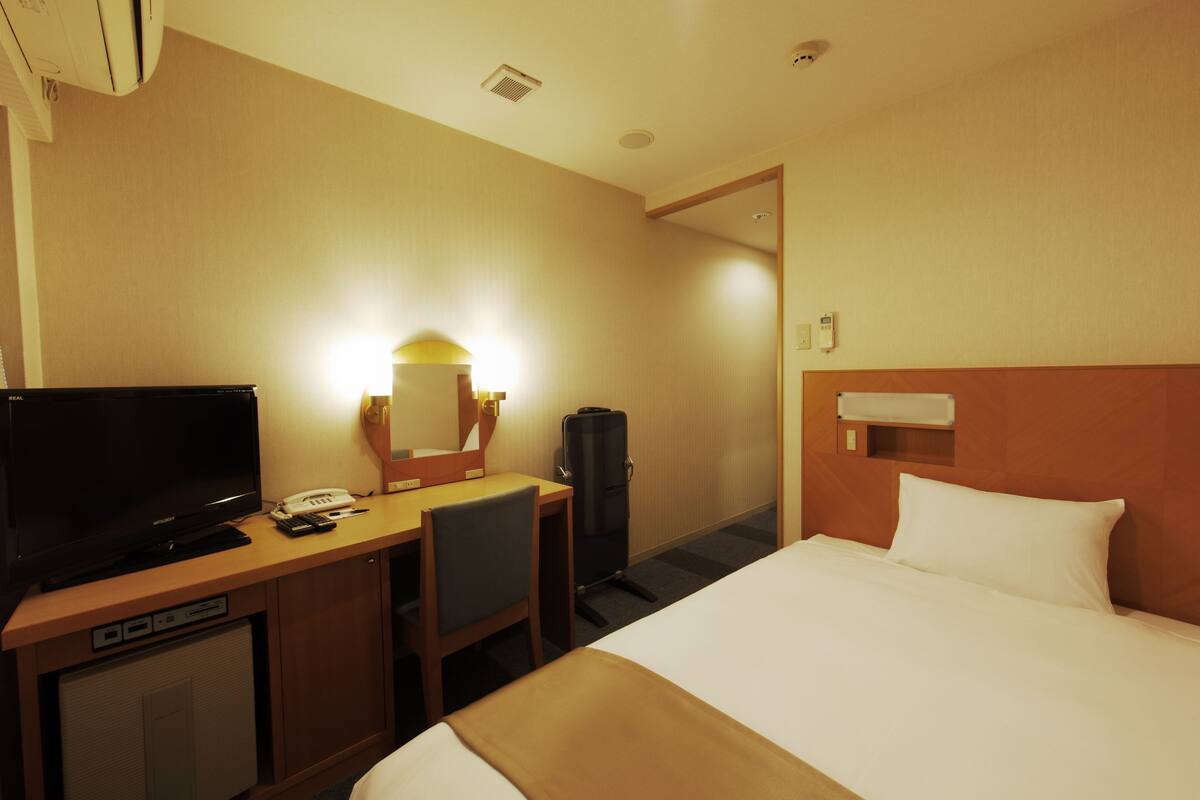 A hotel room with a bed, a desk with a television, a mirror, and a small refrigerator.