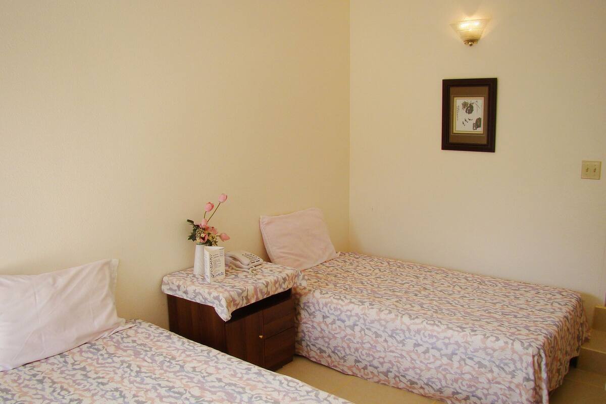 Ein Schlafzimmer mit zwei Betten, einem Nachttisch, einer Blumenvase, einem gerahmten Bild an der Wand und einer Wandleuchte.
