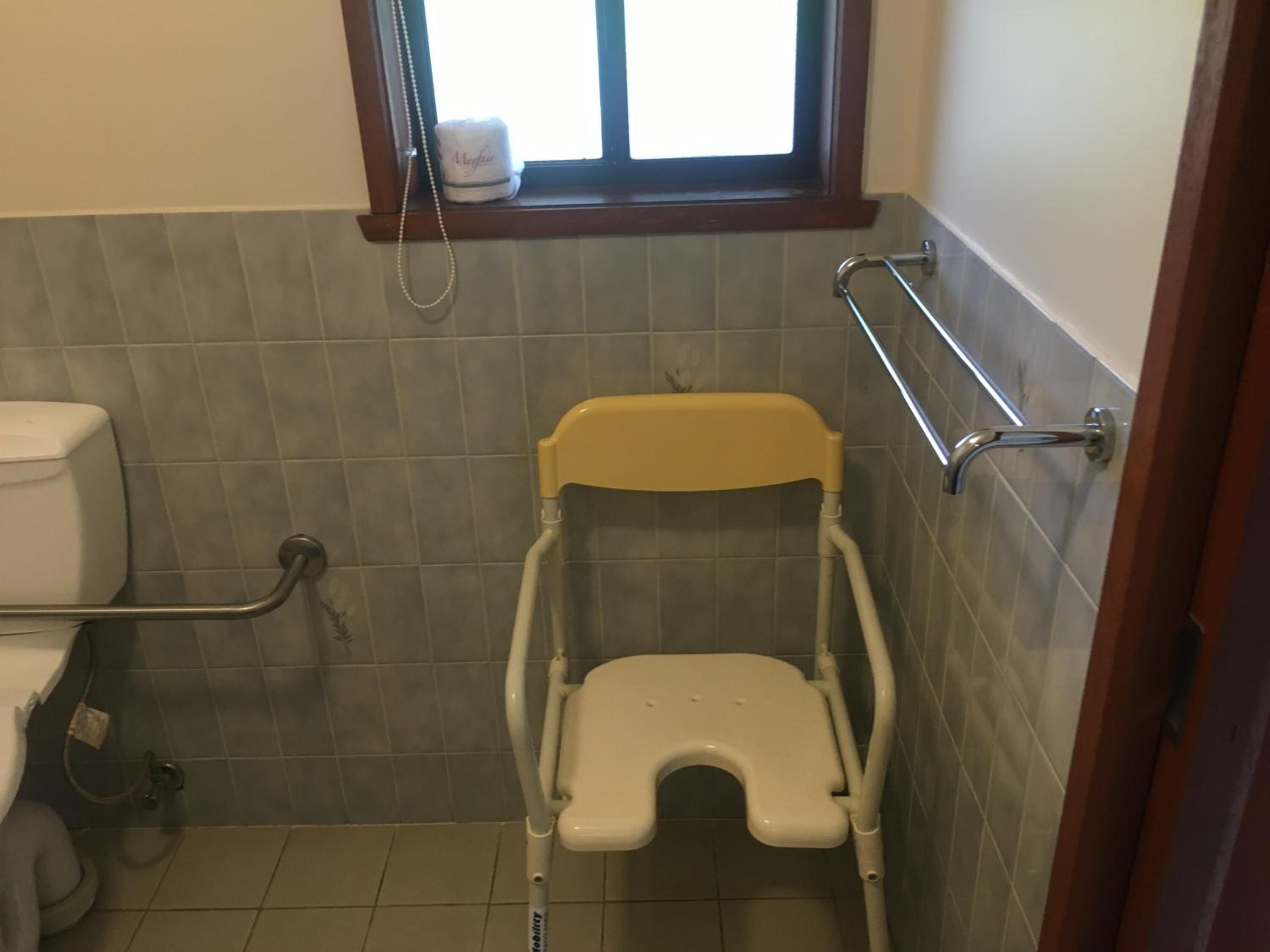 A small bathroom with a toilet, a raised toilet seat, grab bars, and a window with a towel.