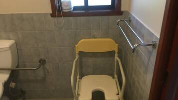 A small bathroom with a toilet, a raised toilet seat, grab bars, and a window with a towel.