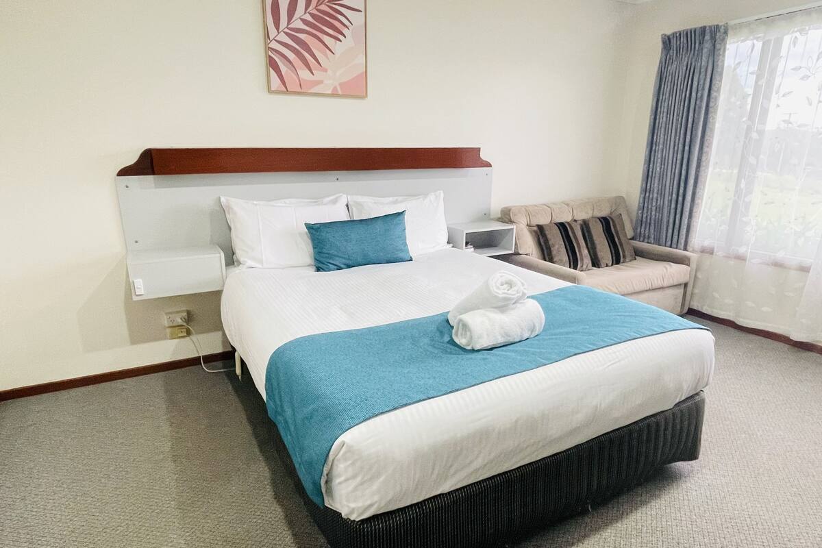 A hotel room with a large bed, a bedside table, a chair, a window with curtains, and a wall-mounted artwork.