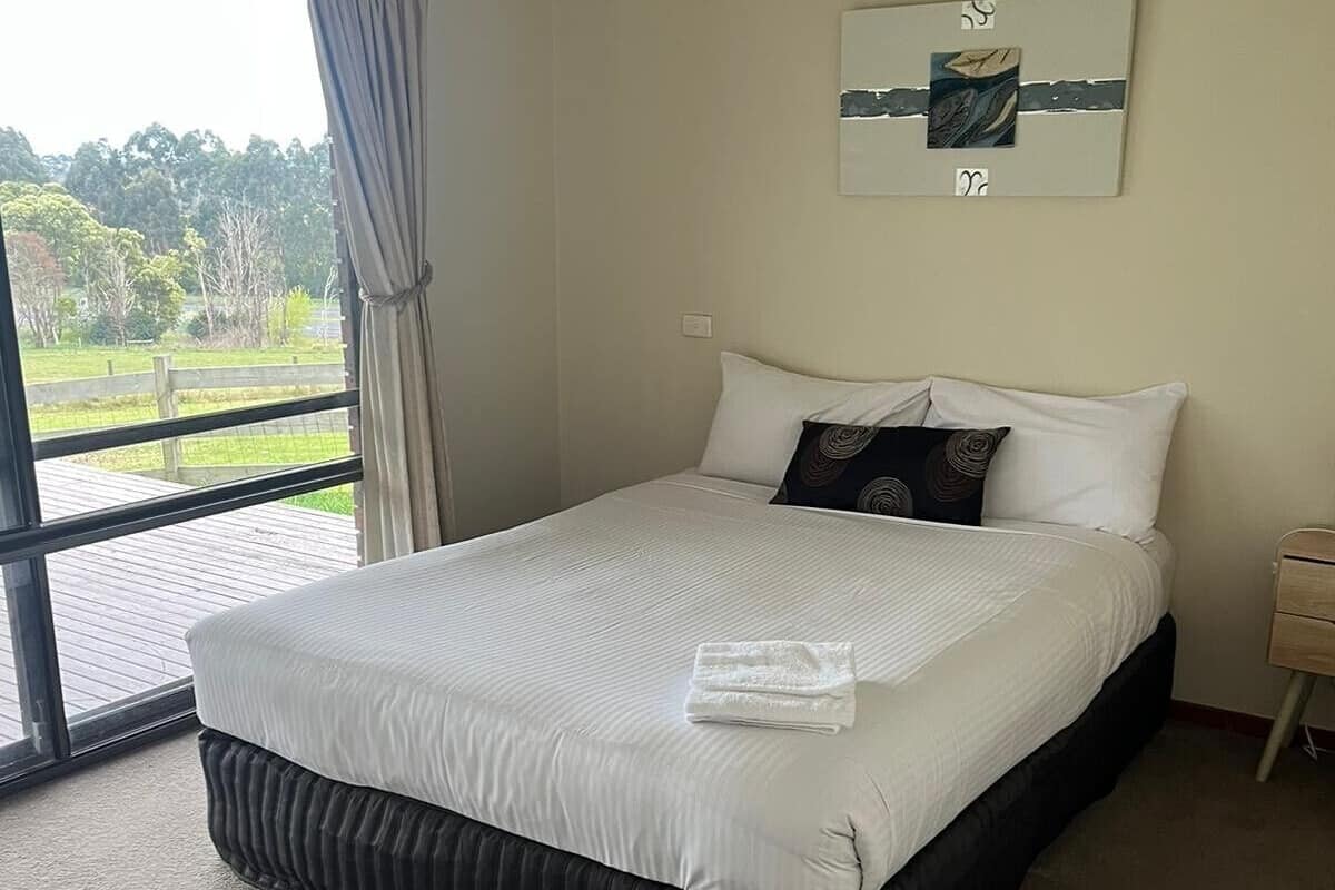 A bedroom with a large bed, a window with curtains, a wall-mounted air conditioner, and a small bedside table.