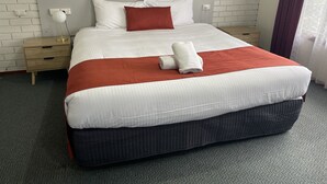 A neatly made bed with white and red bedding, a wooden nightstand with a lamp, and a white brick wall.