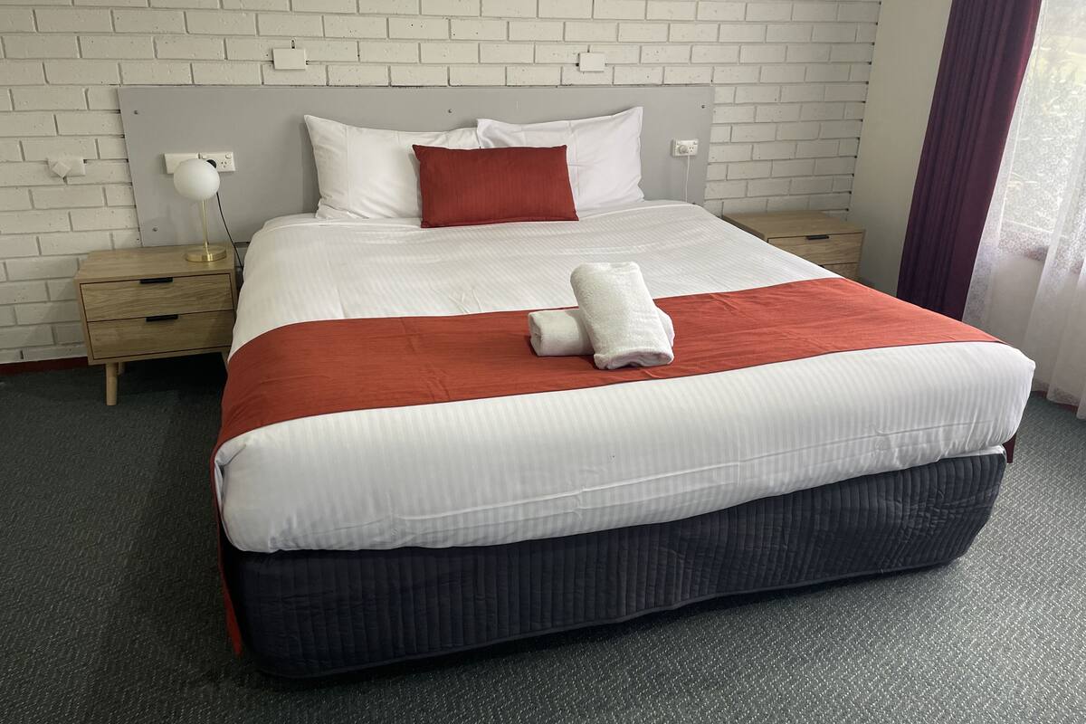 A neatly made bed with white and red bedding, a wooden nightstand with a lamp, and a white brick wall.
