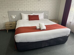 A neatly made bed with white and red bedding, a wooden nightstand with a lamp, and a white brick wall.
