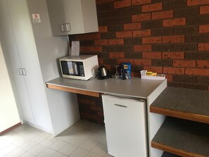 Cocina compacta con microondas, hervidor y refrigerador pequeño. Se observa una pared de ladrillo y armarios blancos.