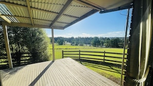 Una terraza de madera con techo metálico, con vistas a un campo verde cercado y árboles.