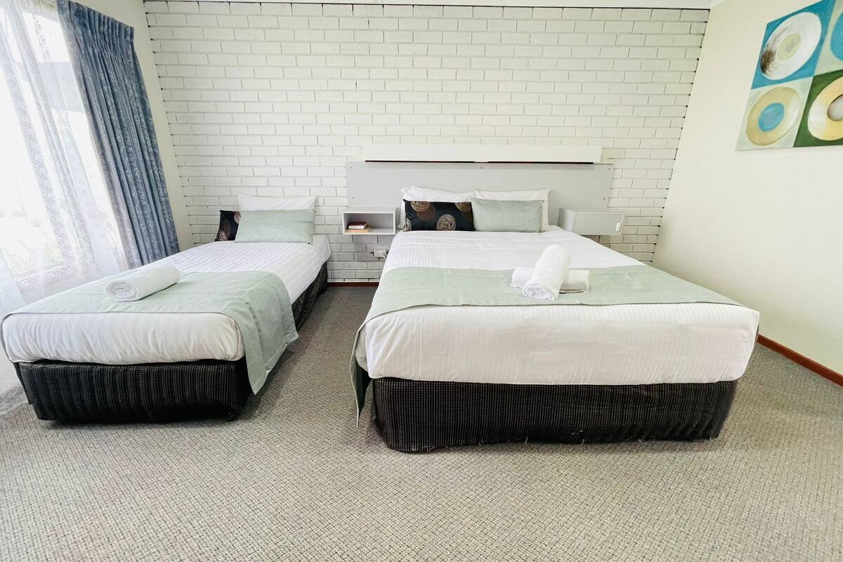 A hotel room with two beds, a white brick wall, a window with curtains, and a painting on the wall.