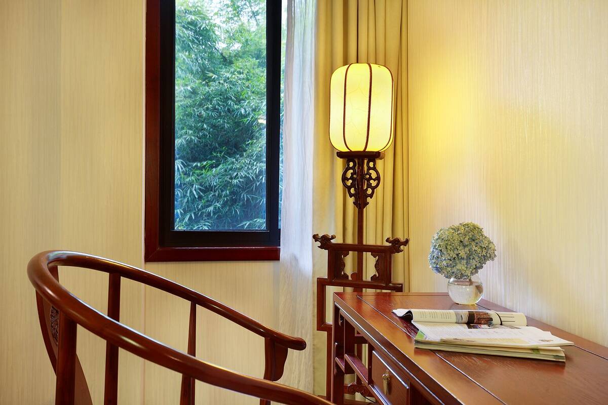 A wooden chair, a desk with papers, a lamp, and a window with a view of greenery.