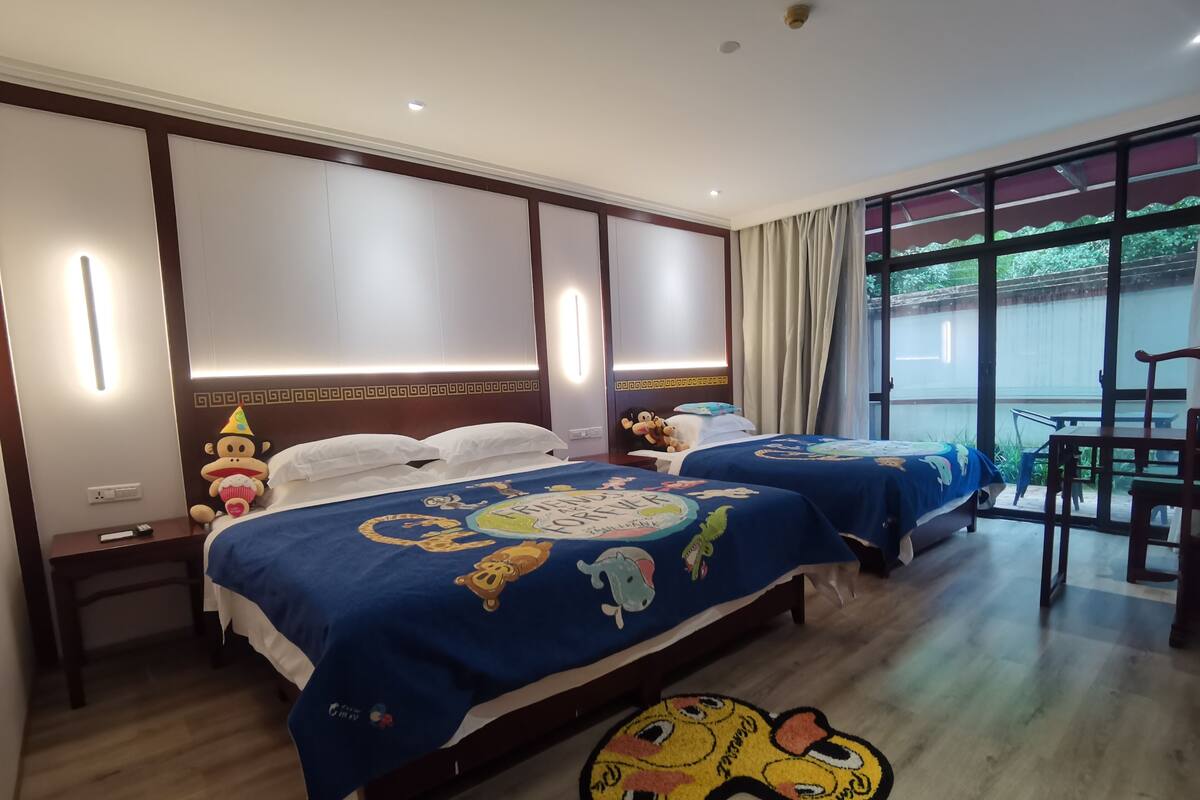 A hotel room with two beds, each with a blue bedspread featuring cartoon characters. There is a small table and a chair in the corner, and a window with curtains.