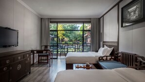 Habitación de hotel con dos camas, un escritorio, una silla, un televisor y un balcón con vistas a zonas verdes y edificios.