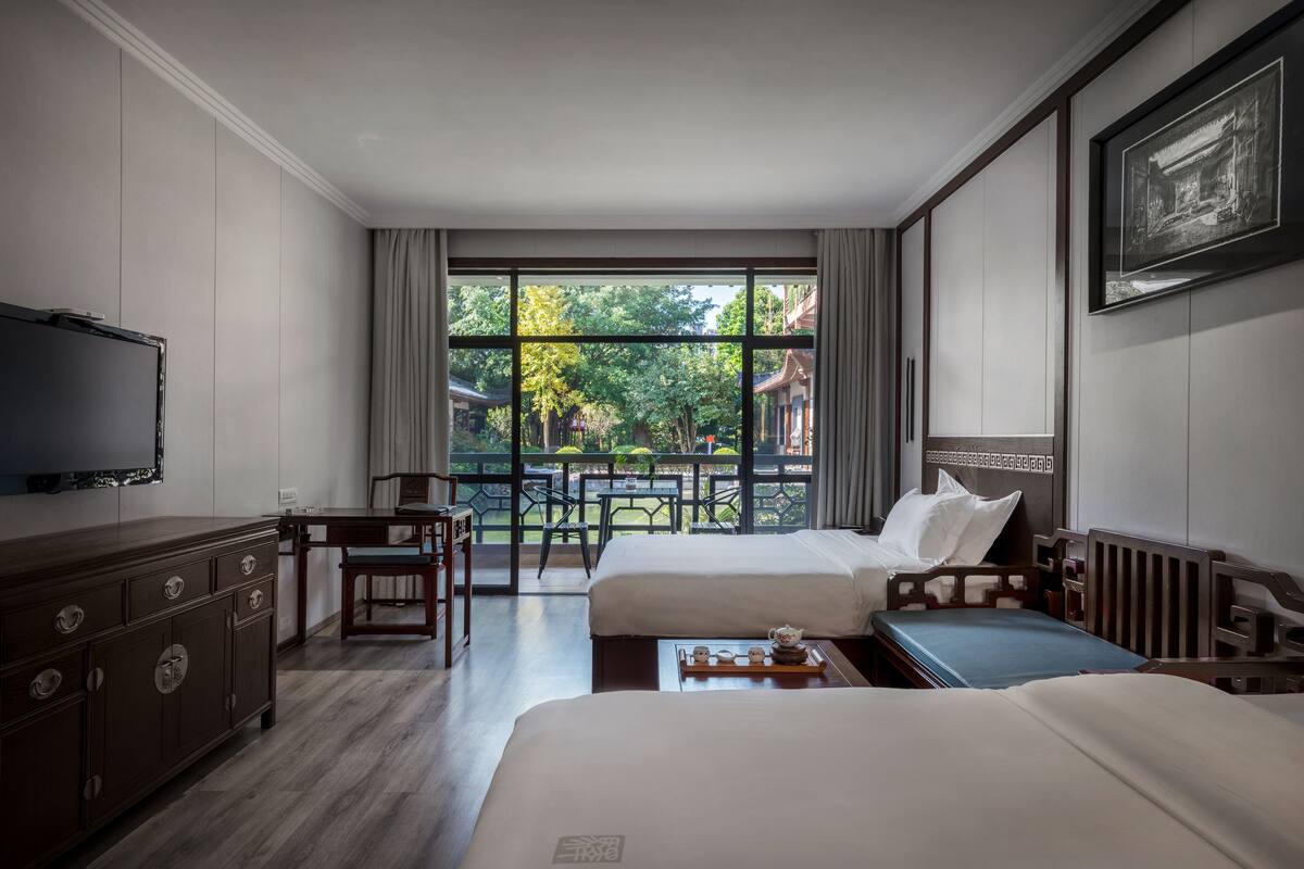 A hotel room with two beds, a desk, a chair, a TV, and a balcony with a view of greenery and buildings.