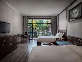 A hotel room with two beds, a desk, a chair, a TV, and a balcony with a view of greenery and buildings.