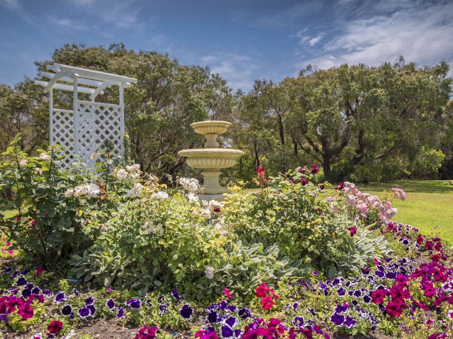 A garden with a white pergola and a tiered fountain surrounded by colorful flowers.